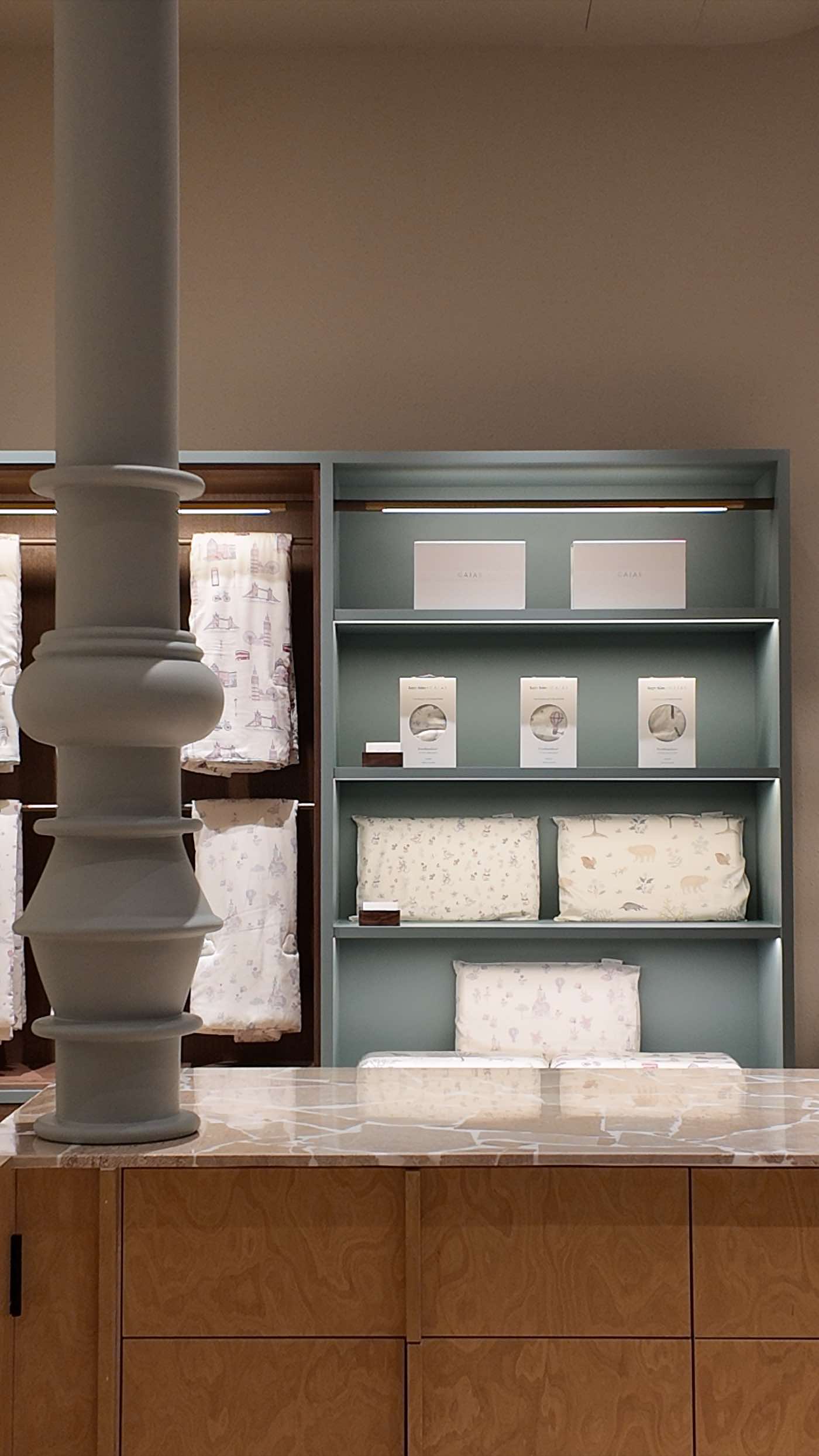 A turned plaster column beside sage-green shelving of children’s bedding